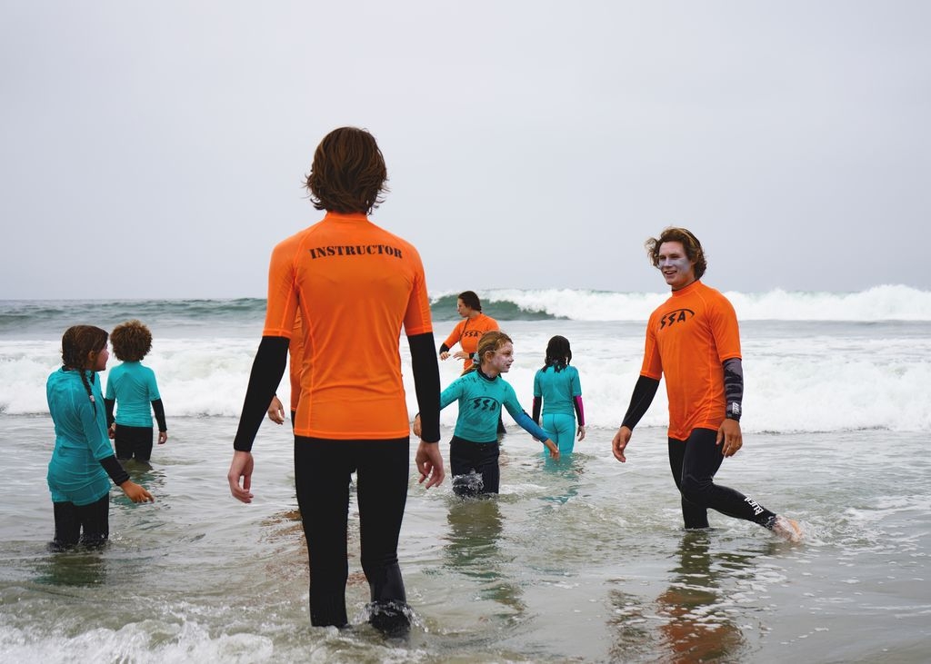 Campers wearing bright blue rashguards are swimming in the ocean in Venice Beach under the supervision of camp counselors wearing bright orange rashguards.