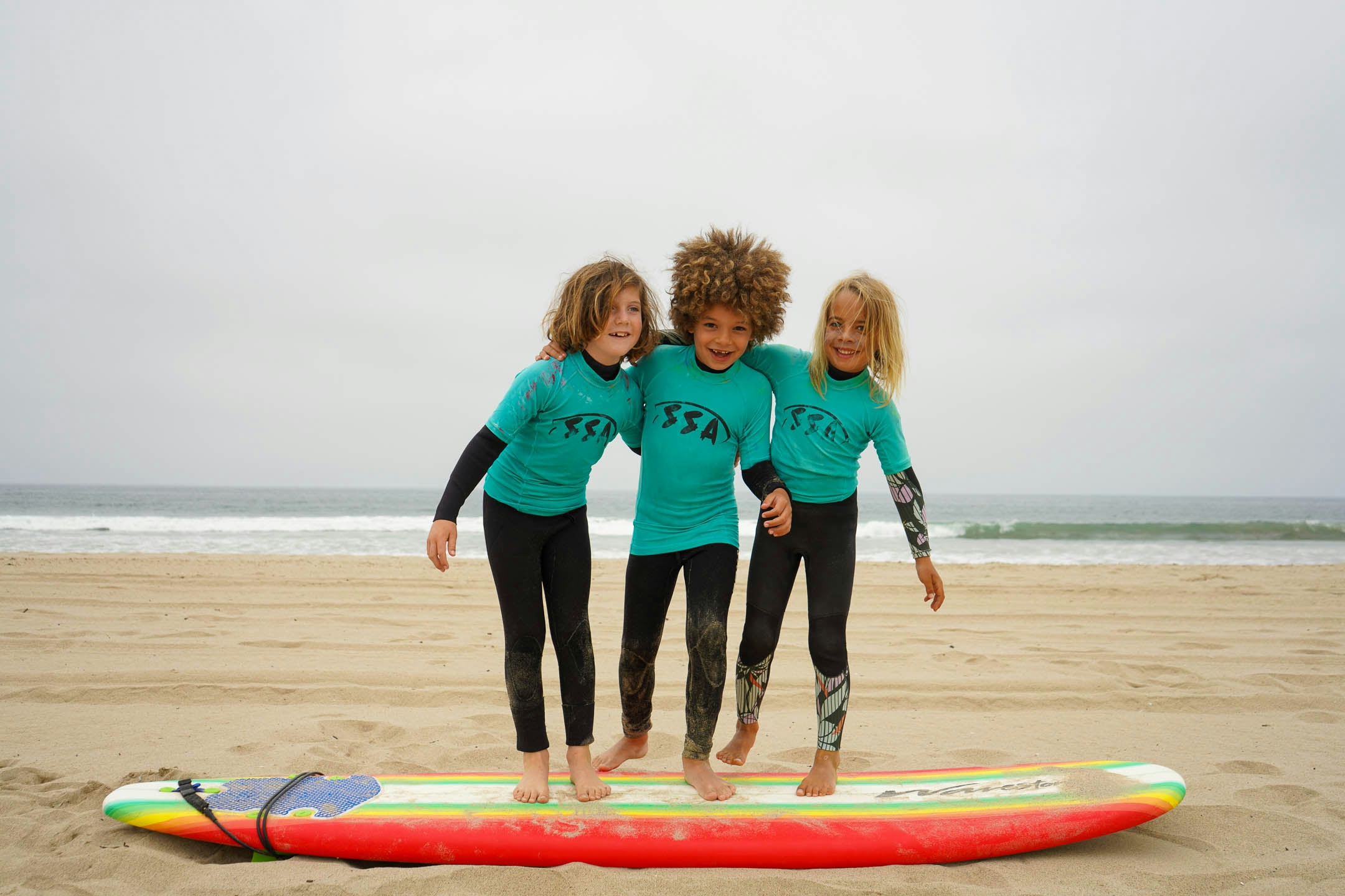 3 campers smiling with their arms around each other. They are standing on a surfboard on the beach with the ocean behind them.